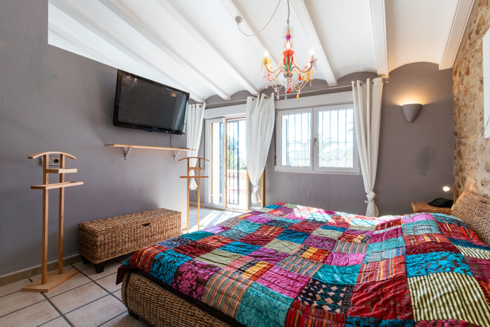 Bedroom featuring a natural stone wall, white beamed ceilings, and a wicker bed frame. It includes a colorful chandelier and direct access to the terrace.