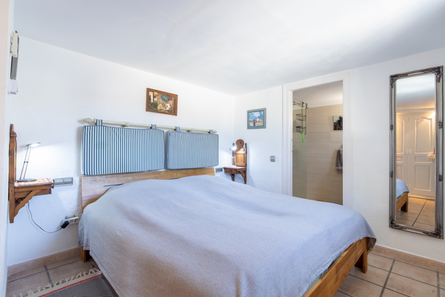 Bedroom featuring a wooden bed frame with striped cushions, wall-mounted nightstands, and terracotta flooring. It includes an en-suite bathroom and a large ornate wall mirror.