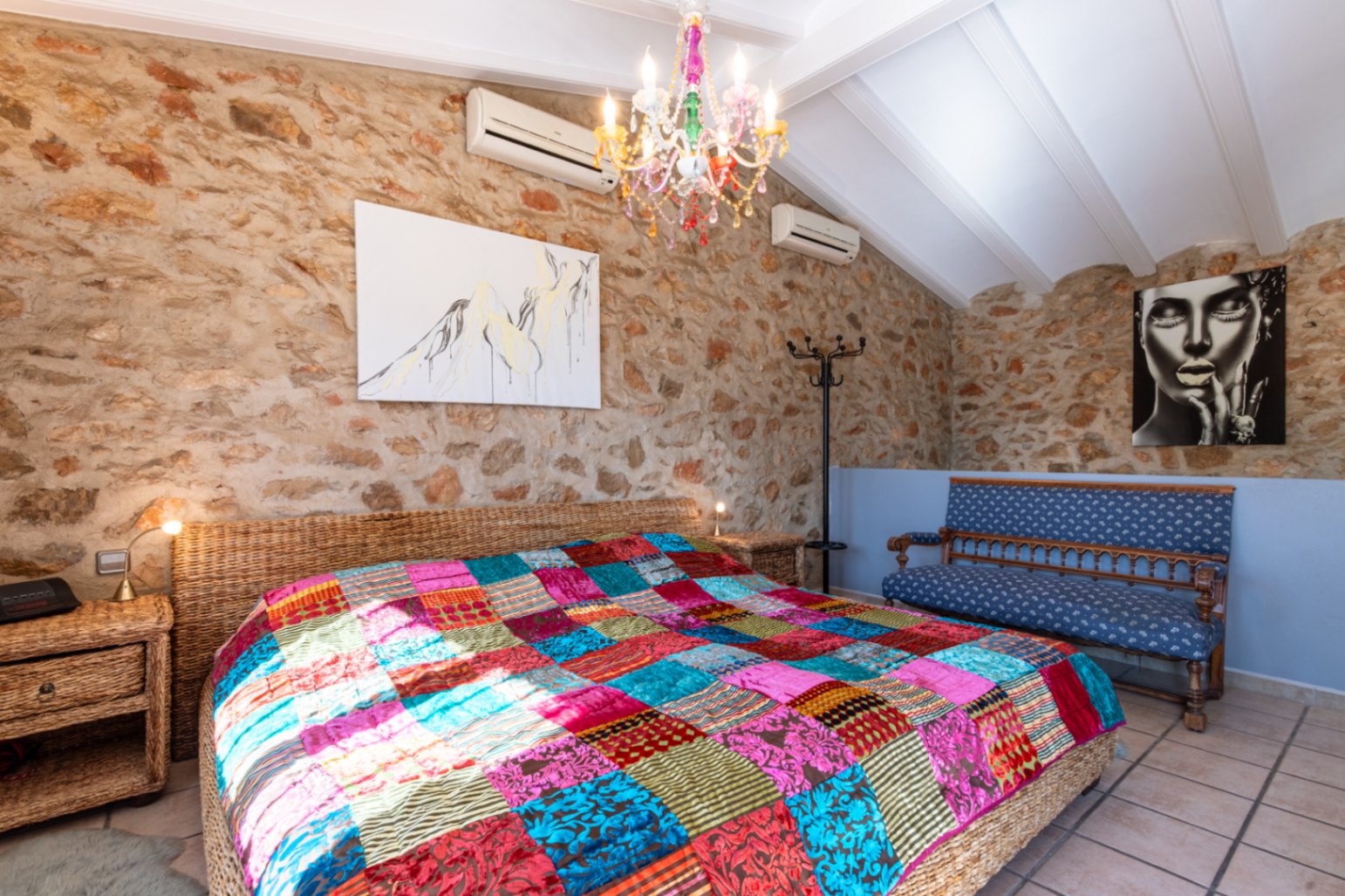 Bedroom featuring exposed natural stone walls, a vaulted beamed ceiling, and a wicker bed frame complemented by a colorful patchwork quilt and a crystal chandelier.