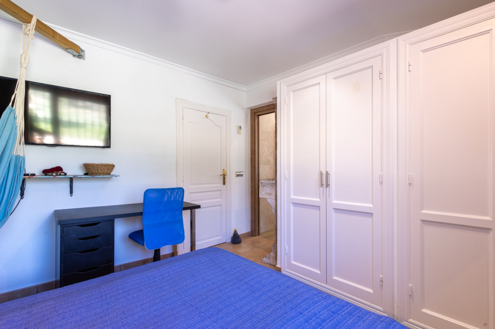Bedroom featuring white built-in wardrobes, a dedicated workspace with a blue chair, and an ensuite bathroom with natural stone details.