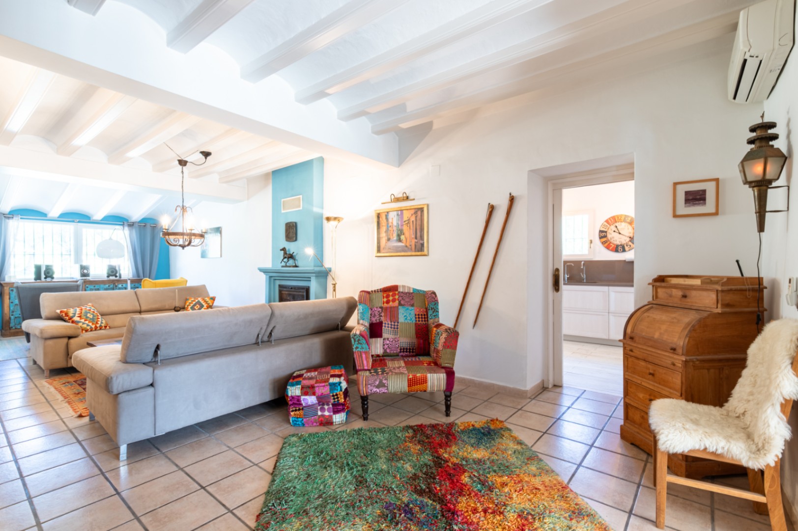 Bright living area featuring white beamed ceilings, a blue accent fireplace, and eclectic decor. The space includes a large sofa and views into the adjacent kitchen.