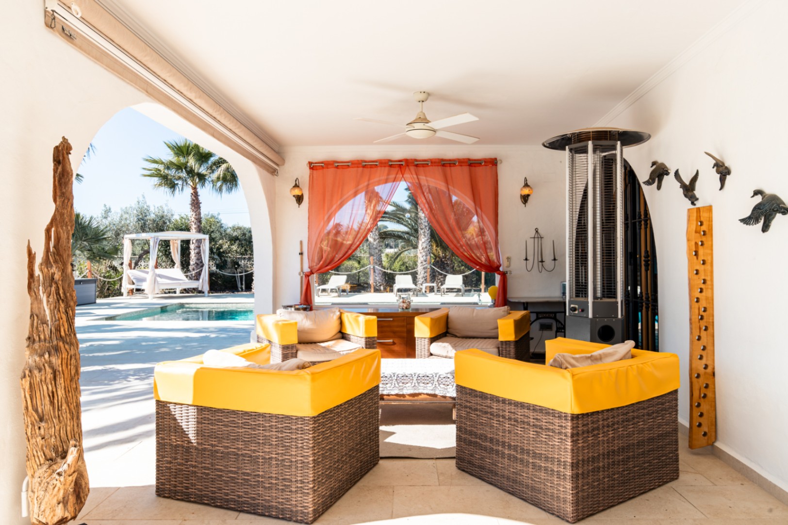 Covered terrace featuring wicker lounge seating with yellow accents, a ceiling fan, and a patio heater, offering views of the pool and landscaped garden.