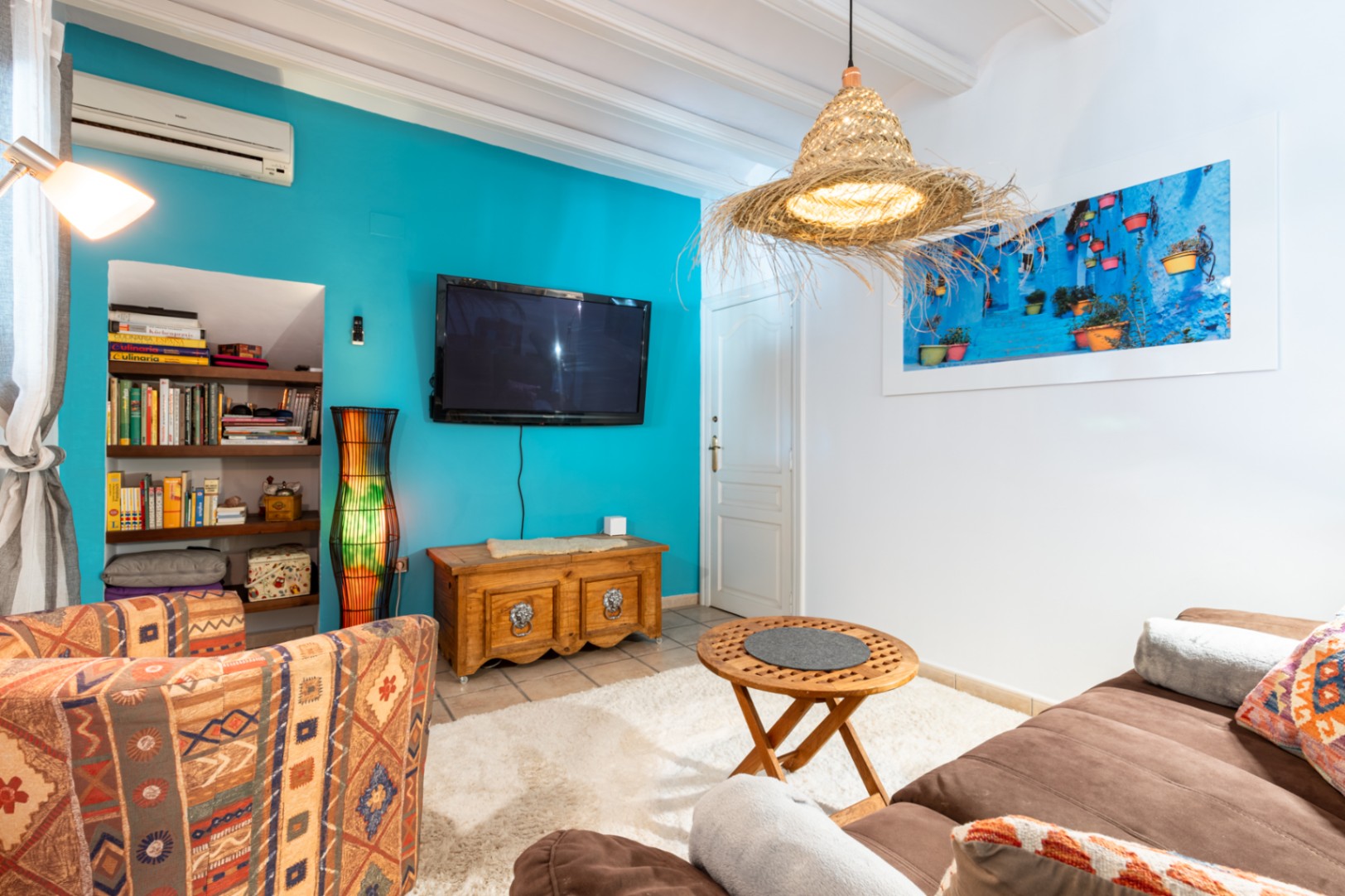 Cozy living space featuring a turquoise accent wall, white beamed ceilings, and built-in shelving. Equipped with air conditioning, rustic wooden furniture, and a natural fiber pendant light.