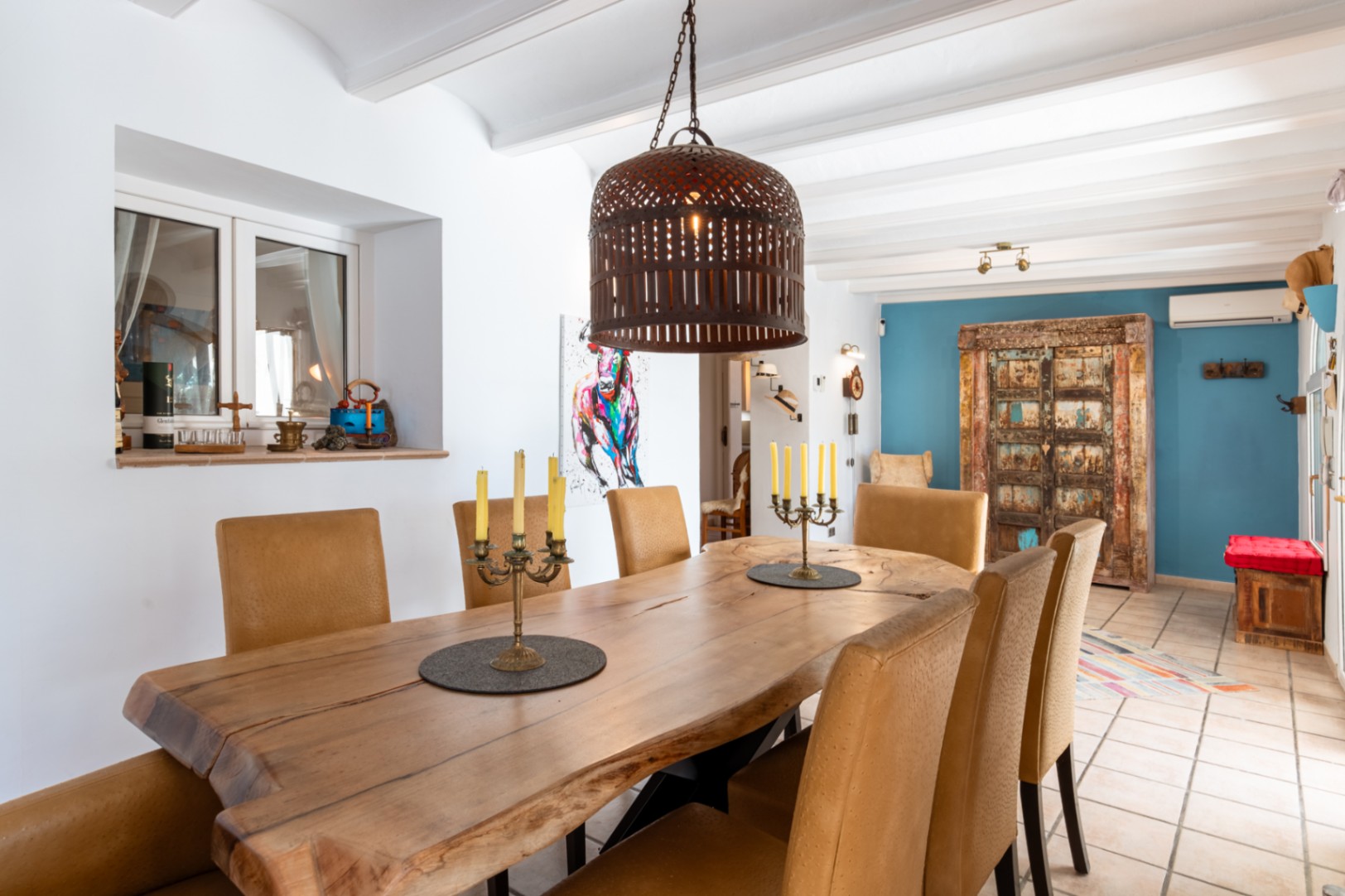 Dining area featuring a live-edge solid wood table, textured chairs, and white beamed ceilings, accented by a blue wall and rustic decorative elements.