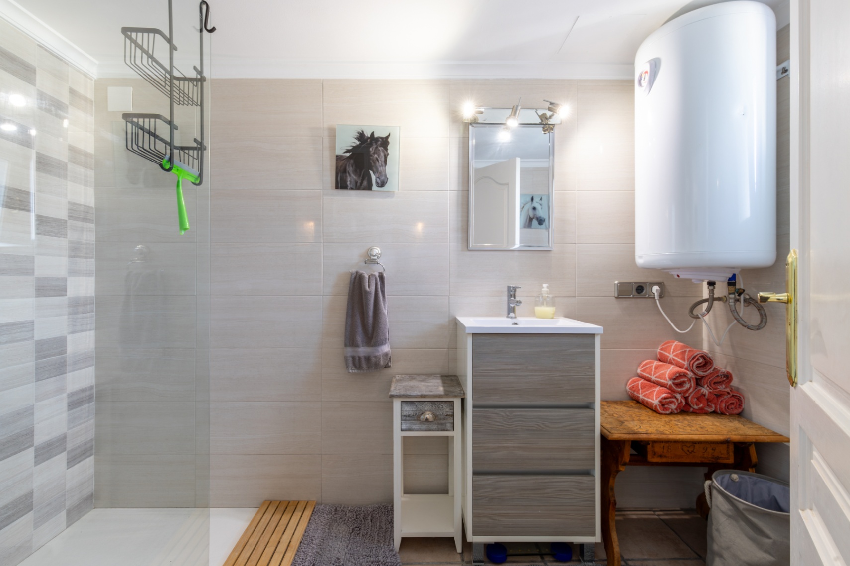 Functional bathroom with a walk-in shower, glass screen, and textured tiling. It features a grey vanity unit, an electric water heater, and rustic wooden furniture.
