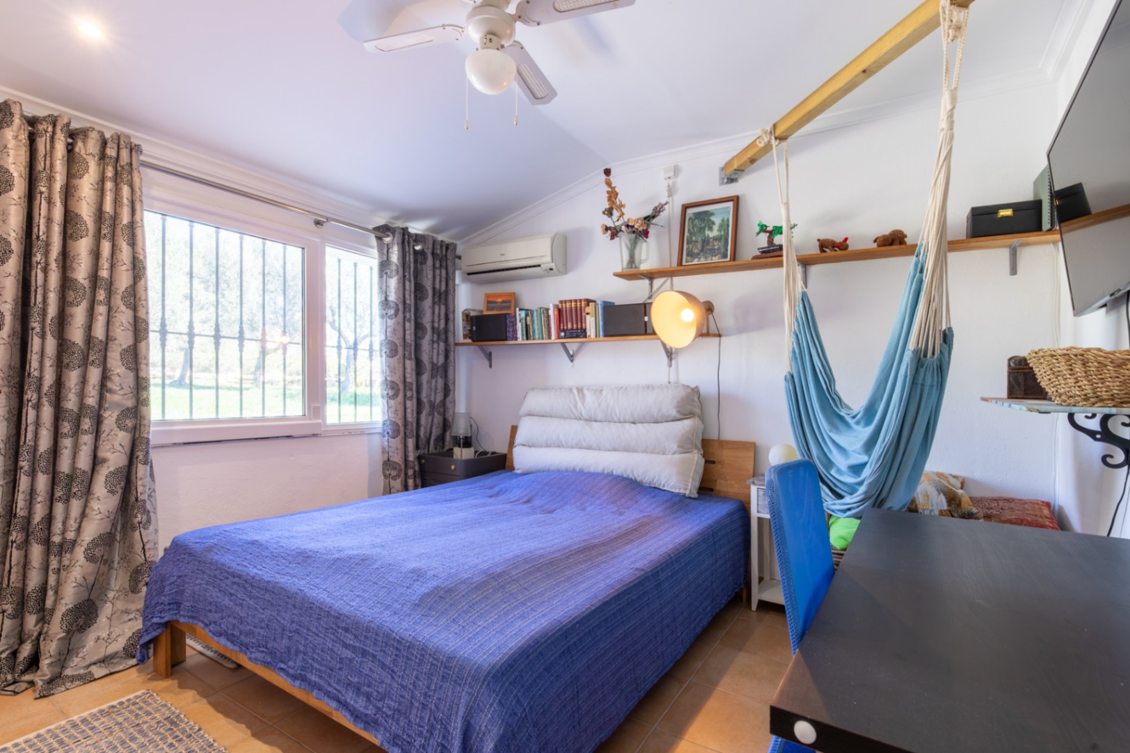 Functional bedroom with a double bed, desk area, wooden shelves, and a hanging hammock. Features include split air conditioning, a ceiling fan, and ample natural light.