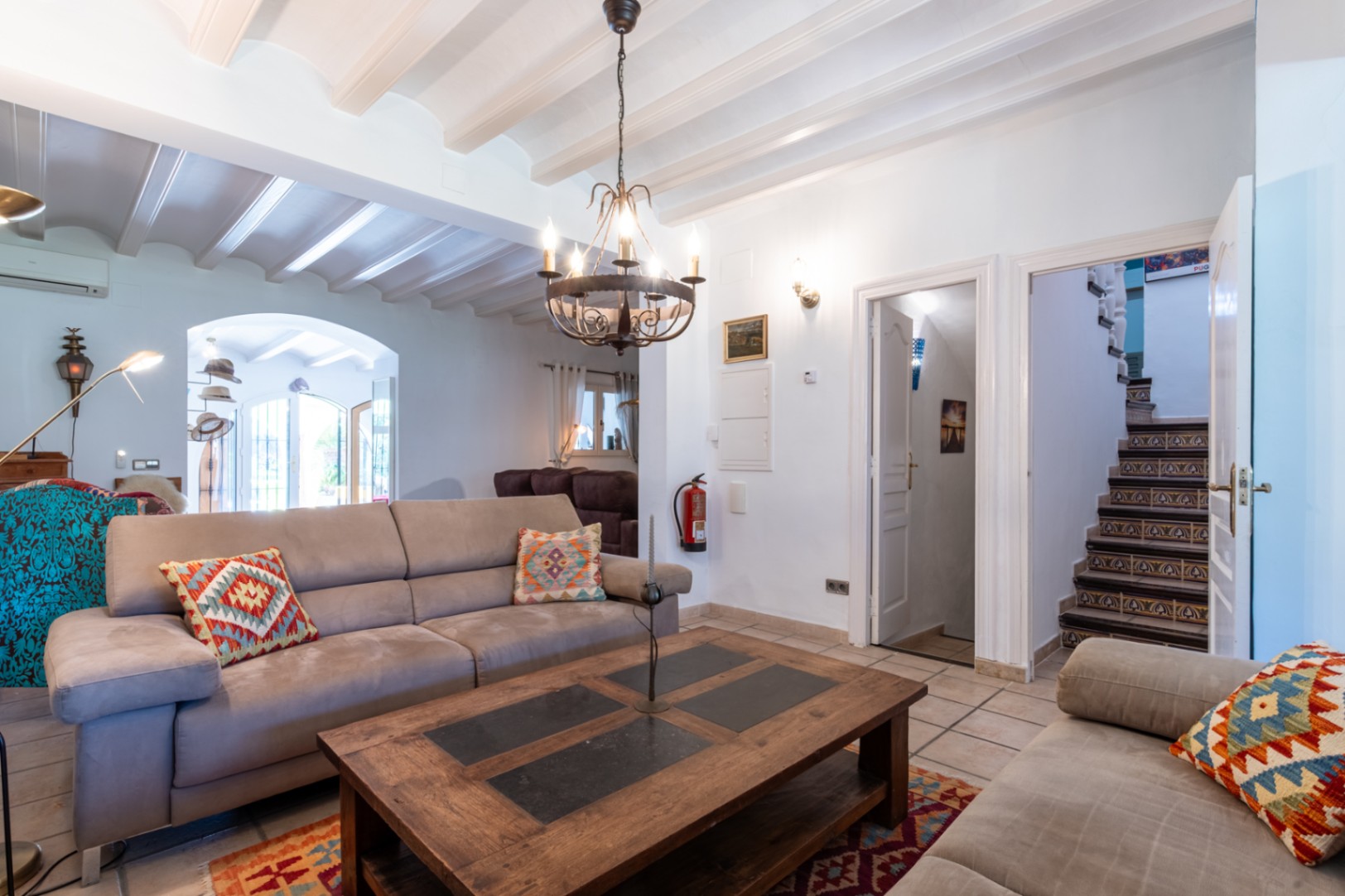 Living area featuring white beamed ceilings, rustic tiled floors, and a staircase with decorative risers. A solid wood coffee table sits under a wrought iron chandelier.