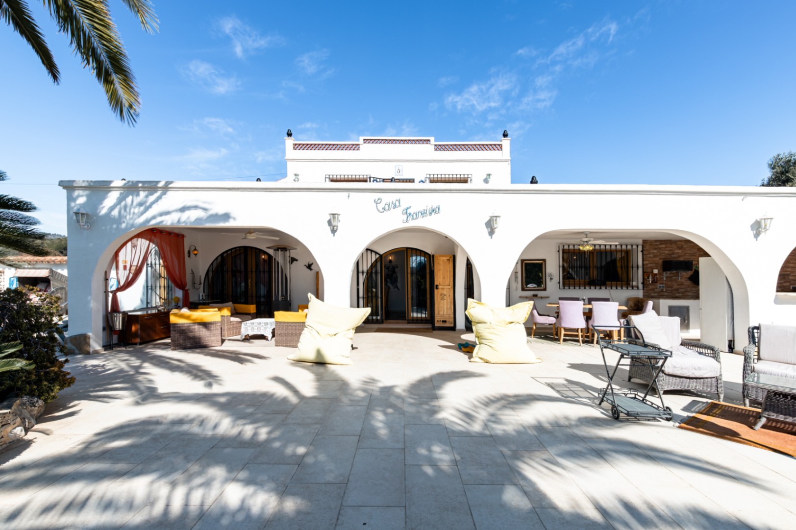 Mediterranean-style white facade featuring an arched naya, stone-paved terrace, and outdoor lounge areas beneath the "Casa Franziska" sign.