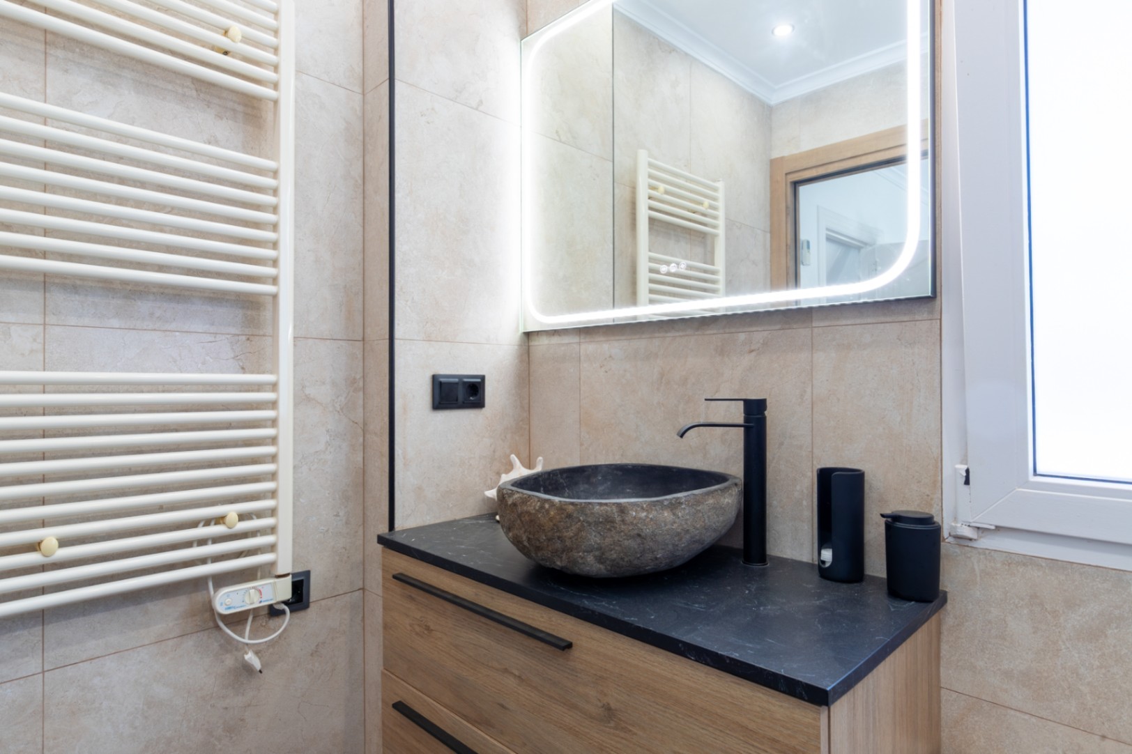 Modern bathroom featuring a natural stone vessel sink on a black countertop with a wooden vanity. Includes a backlit LED mirror and a white heated towel rail.