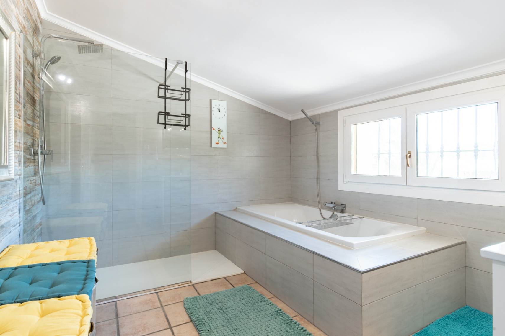 Modern bathroom featuring a walk-in shower with glass partition and a built-in bathtub. It includes grey wall tiling, chrome fixtures, and natural light from a window.