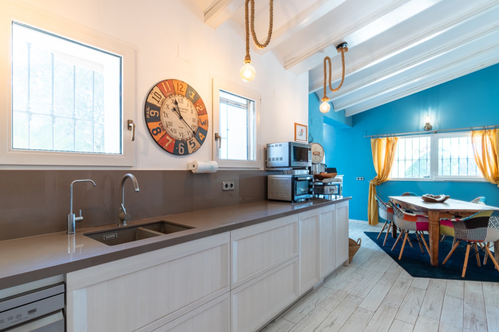 Modern kitchen featuring quartz countertops and integrated sink, opening into a bright dining area with blue accent walls, exposed white beams, and rustic rope lighting.