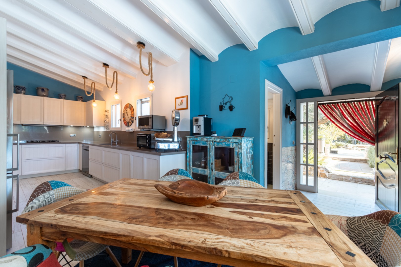 Modern kitchen featuring white beamed ceilings, blue accent walls, a rustic wooden table, and rope pendant lighting. Includes high-end appliances and outdoor access.