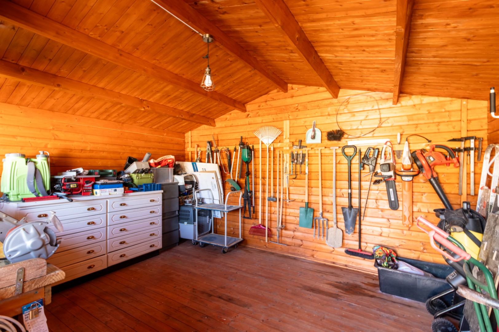 Organized wooden garden shed featuring wall-mounted tool storage, a white chest of drawers, and exposed timber beams.