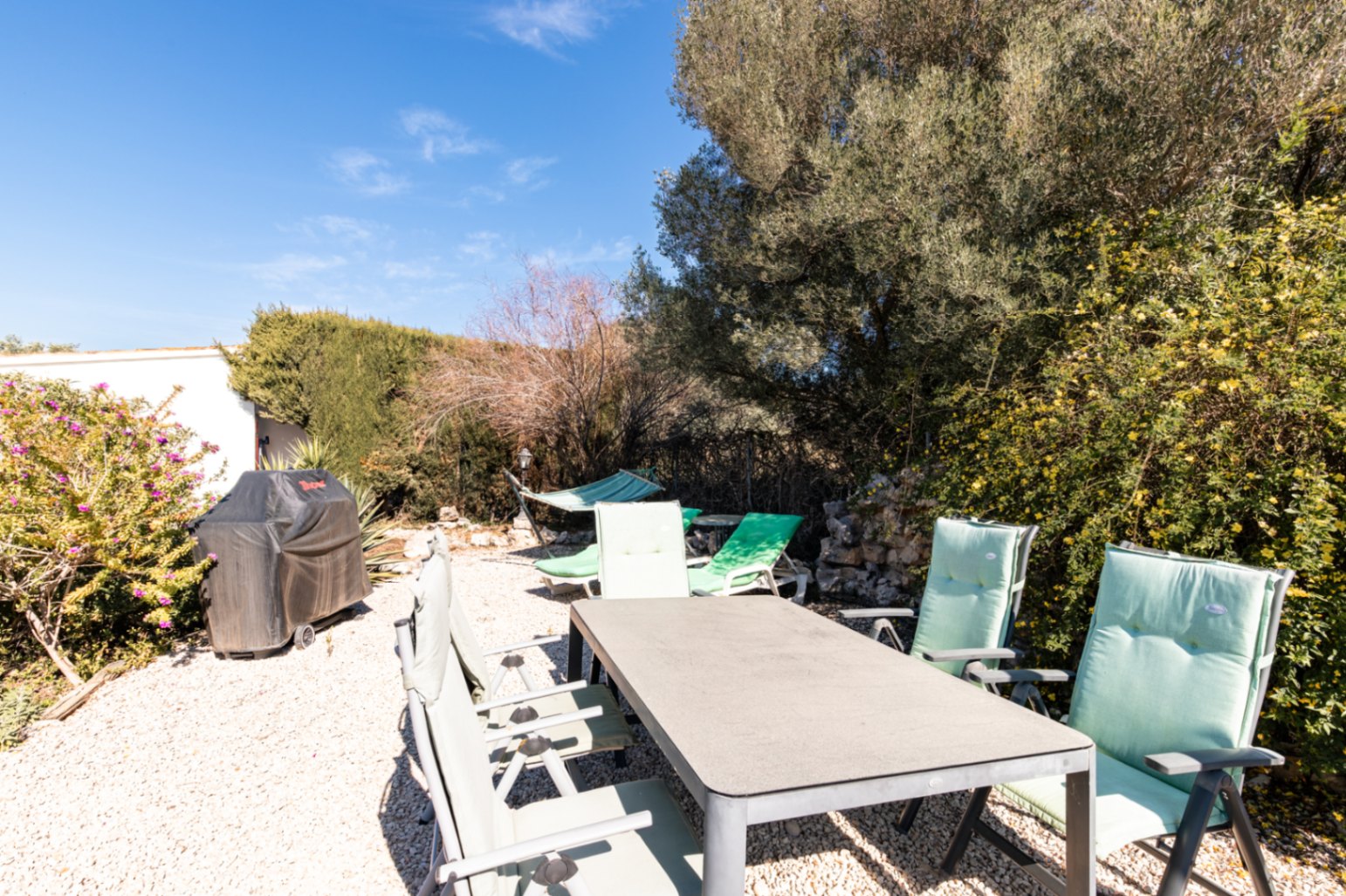 Outdoor dining space set on gravel featuring a rectangular table, cushioned seating, a covered grill, and a hammock amongst mature Mediterranean trees.