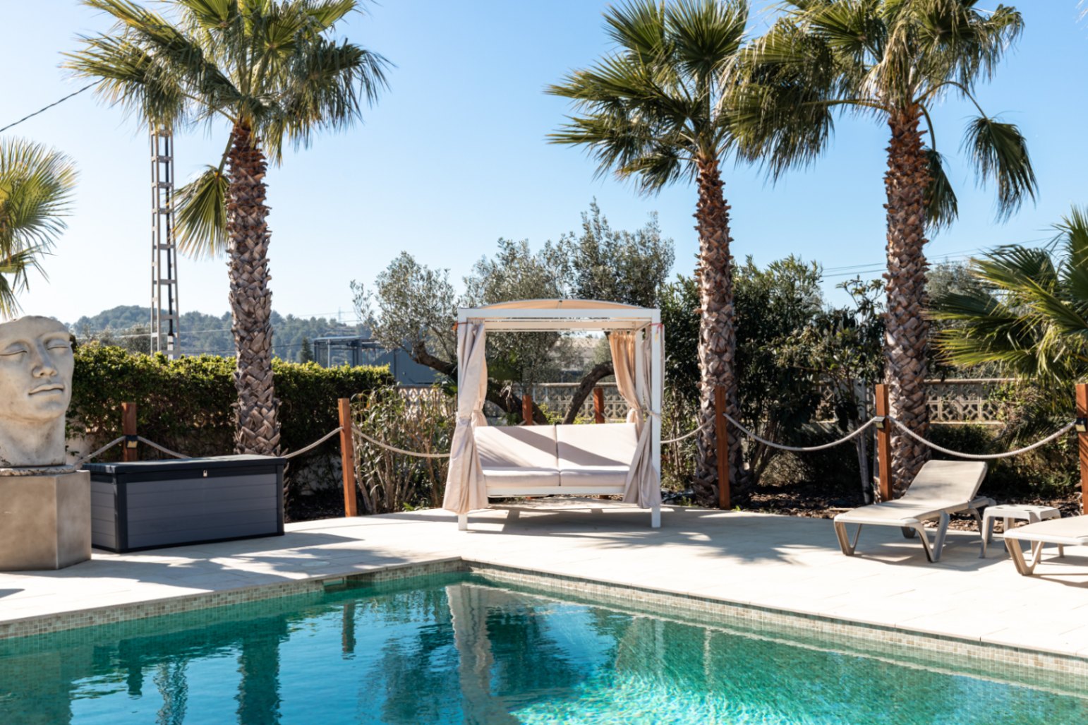 Poolside area featuring a stone terrace, a white Balinese daybed with curtains, sun loungers, and mature palm trees under a clear blue sky.