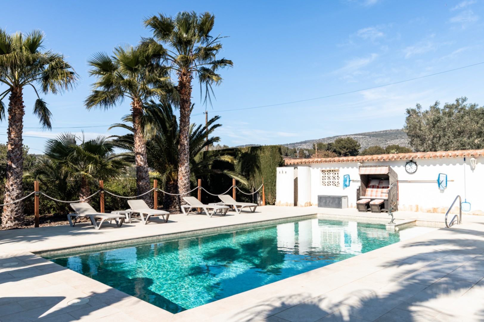 Private 9.5m x 5m swimming pool featuring a light stone terrace, sun loungers, and mature palm trees, complemented by a traditional beach chair and mountain views.