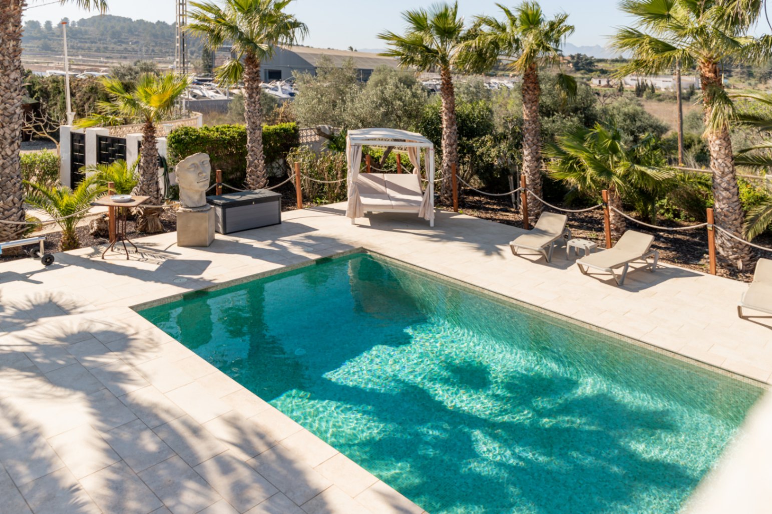 Rectangular swimming pool featuring light stone paving, sun loungers, and a canopy daybed, framed by mature palm trees and decorative garden elements.