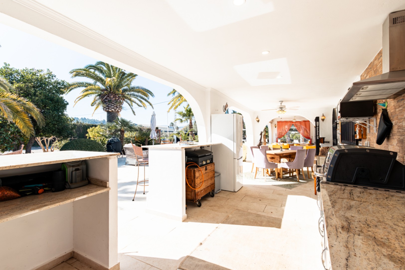 Spacious covered naya featuring a fully equipped outdoor kitchen with stone accents, dining area, and arched views over the palm-lined garden.