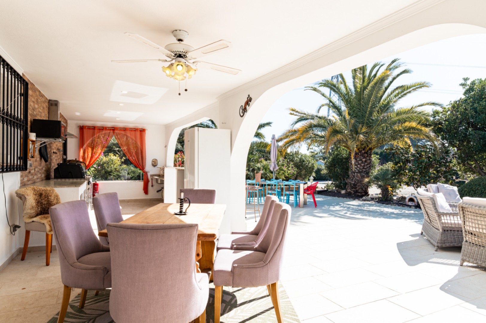 Spacious covered naya featuring an outdoor dining area with a ceiling fan and a summer kitchen. The terrace overlooks a sunny garden with mature palm trees.