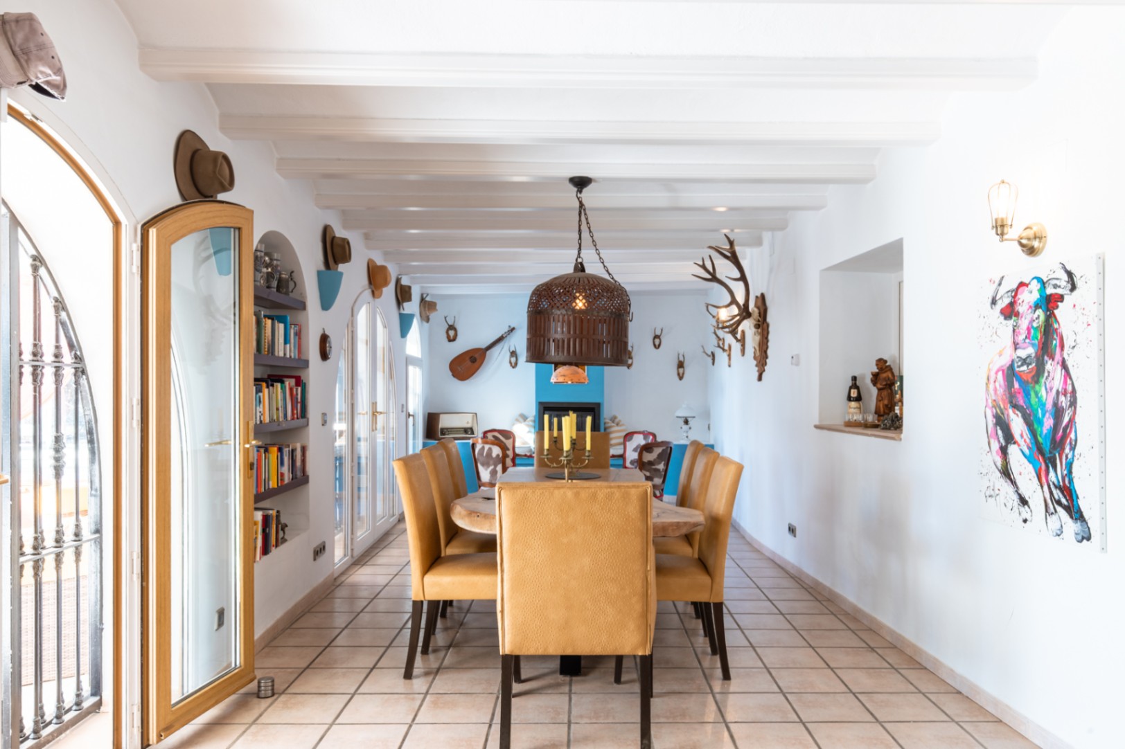 Spacious dining room featuring white exposed beams, a solid wood table, and mustard upholstered chairs. The area includes arched glass doors, built-in shelving, and rustic wall decor.