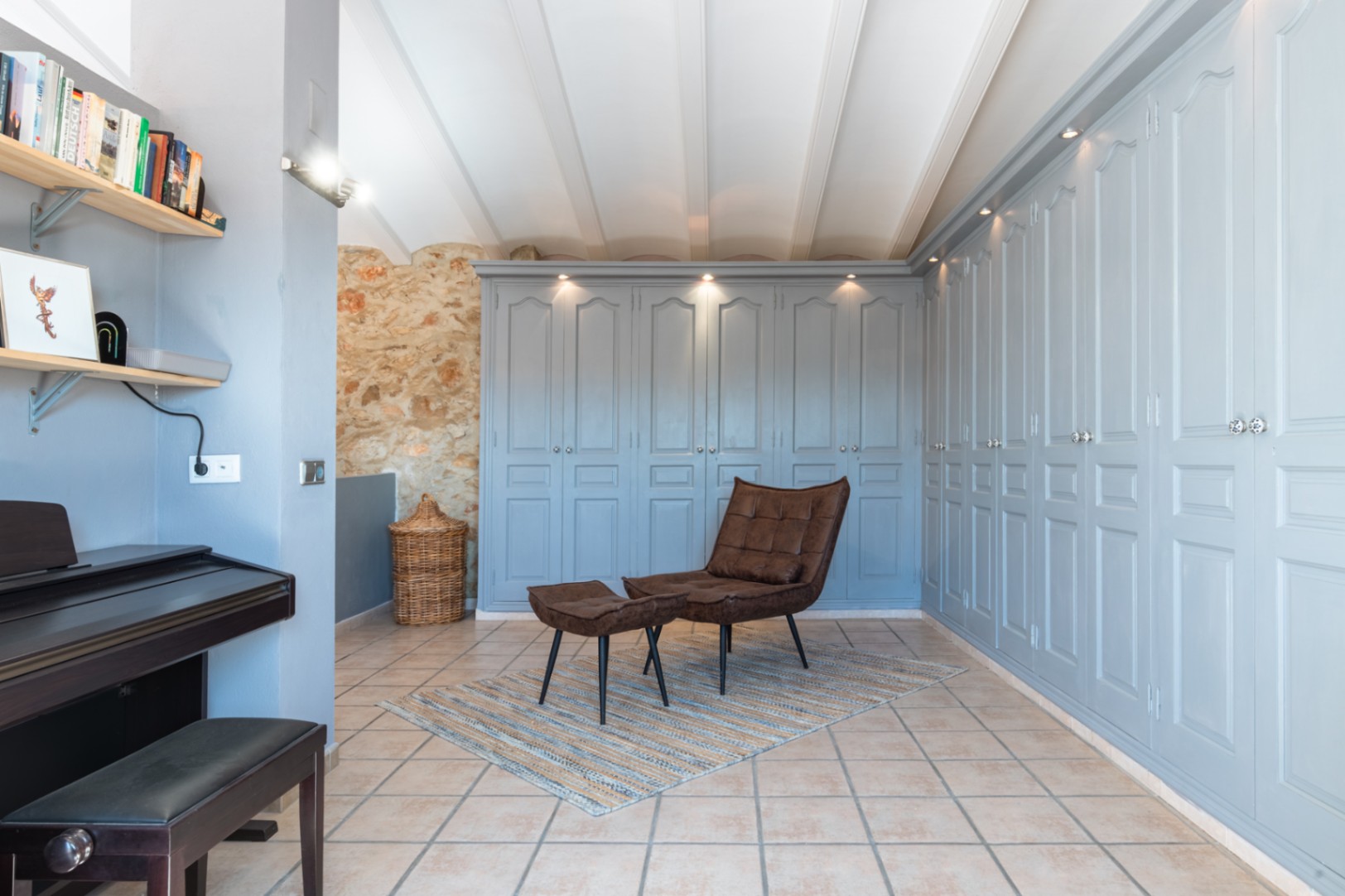 Transitional space featuring extensive built-in wardrobes in soft blue, exposed stone masonry, white beamed ceilings, and a relaxation area with a leather lounge chair and piano.