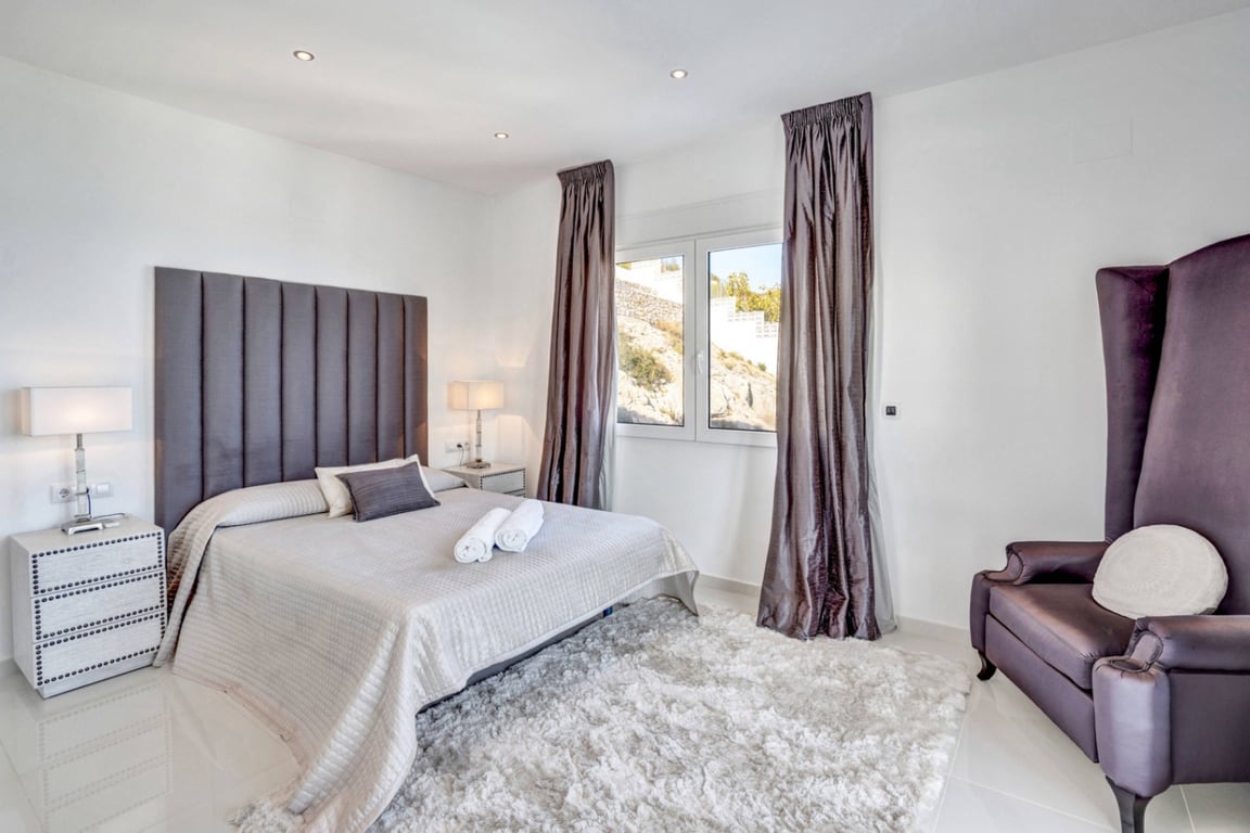 Bedroom suite featuring a tall grey headboard, plush white rug, and a distinctive high-back armchair. The space includes recessed lighting and views of the rocky landscape.
