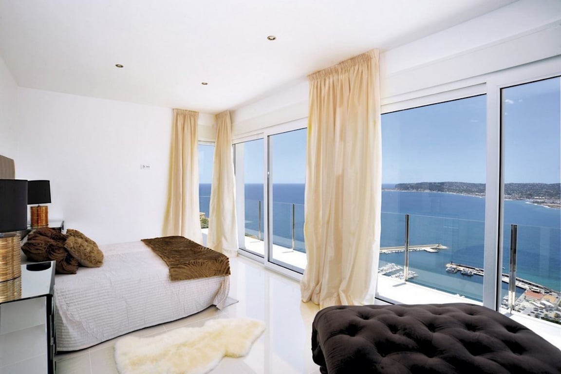 Bedroom suite with floor-to-ceiling windows offering panoramic views of Jávea port and the sea. Features polished floors, recessed lighting, and direct terrace access.