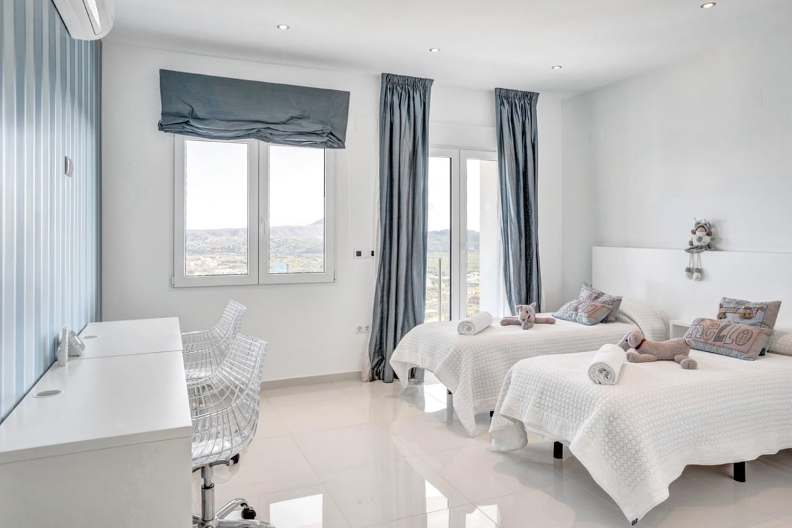 Bedroom with twin beds, a white desk with designer chairs, and large windows showcasing panoramic mountain and sea views in Jávea.