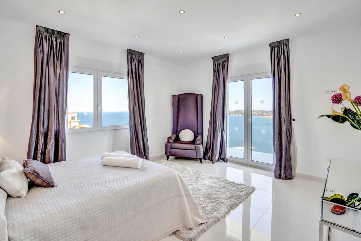 Bright bedroom featuring a purple velvet wingback chair, tiled floors, and large windows offering expansive views of the Mediterranean coastline.