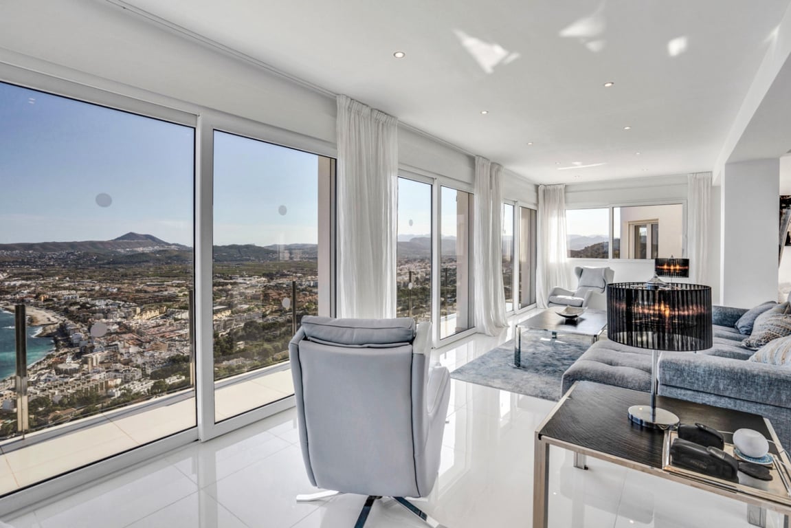 Bright living area featuring floor-to-ceiling windows with panoramic views of Jávea bay and mountains, complemented by polished white flooring and recessed lighting.