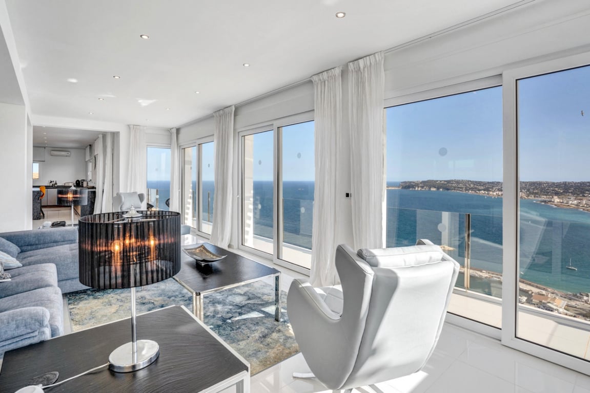 Bright living room featuring floor-to-ceiling sliding doors with panoramic sea views. The space includes modern sofas, a designer armchair, and recessed ceiling lighting.