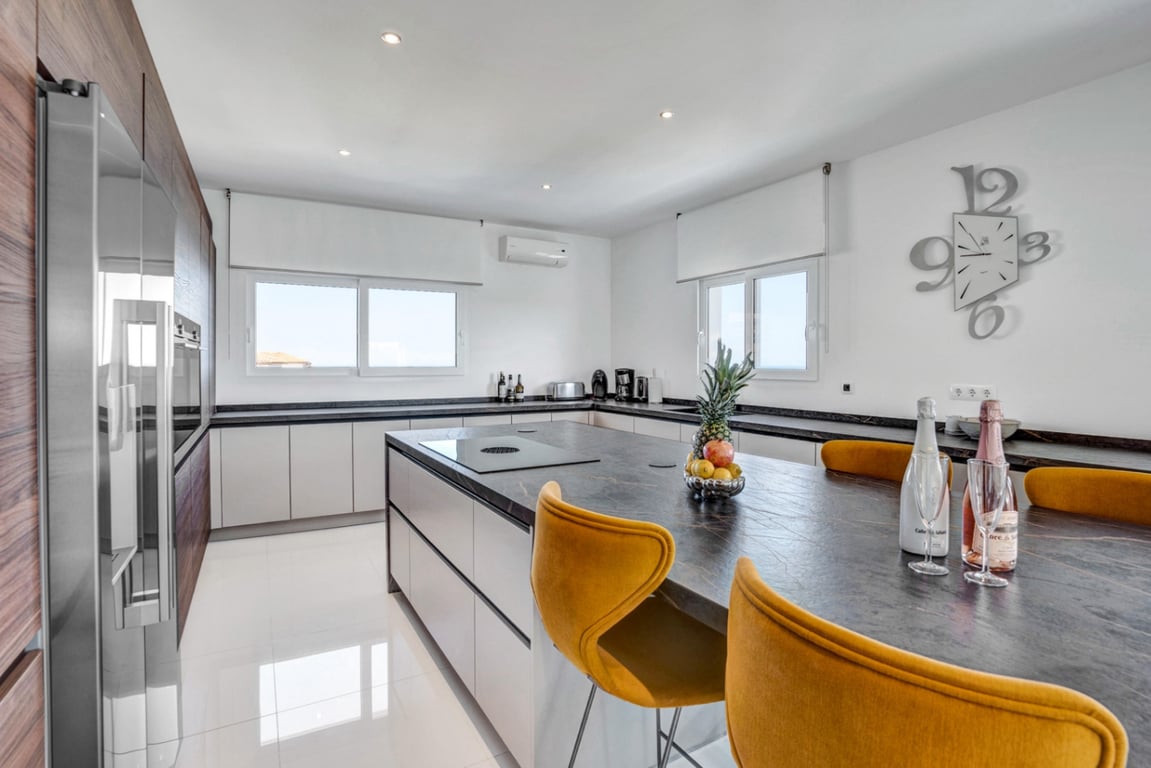 Designer kitchen featuring a dark stone island with integrated hob, mustard yellow bar stools, and sleek wood-paneled cabinetry on high-gloss floors.