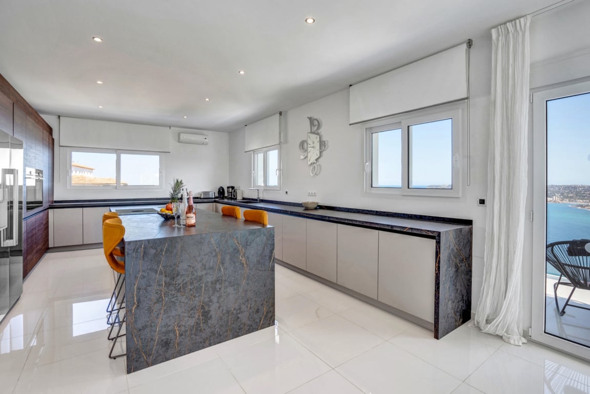 Designer kitchen with a veined stone island, mustard bar stools, and integrated appliances. Large windows and glass doors frame panoramic views of the coast.