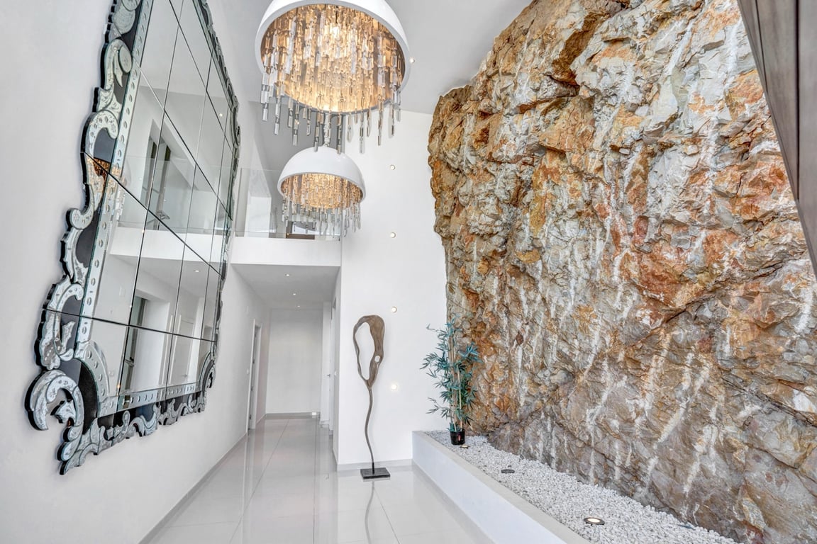 Double-height hallway featuring an integrated natural rock wall, designer crystal chandeliers, and a large ornate mirror against polished flooring.