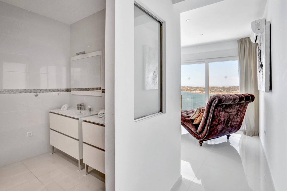 En-suite bathroom featuring a double vanity and mosaic tile accents, opening onto a bright seating area with a velvet chaise longue and Mediterranean views.