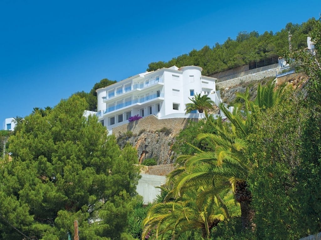 Exterior view of the white multi-level villa built into the hillside, featuring glass balconies, a rounded turret, and lush Mediterranean vegetation.