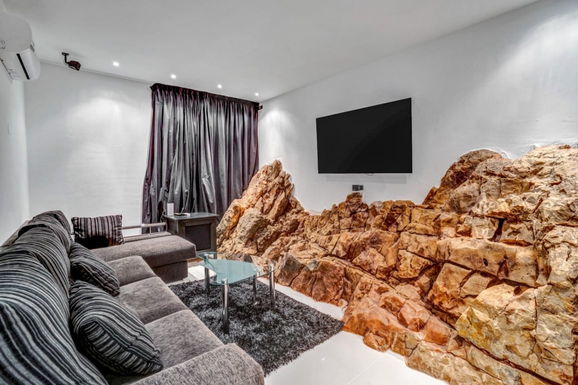 Home cinema room featuring natural rock integrated into the wall, a grey sofa, glass coffee table, and wall-mounted TV with recessed lighting.