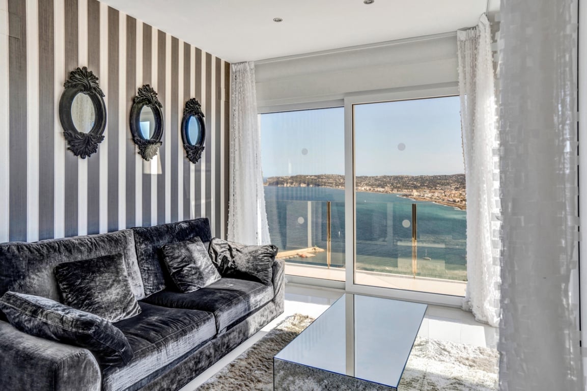 Living area featuring a grey velvet sofa, striped wallpaper, and ornate mirrors, with floor-to-ceiling windows overlooking the Mediterranean coast.