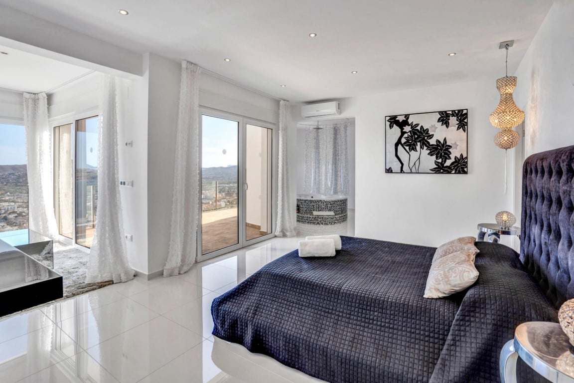 Master suite featuring a tufted headboard, polished tile floors, and large glass doors. An integrated mosaic jacuzzi offers views over the bay of Jávea.