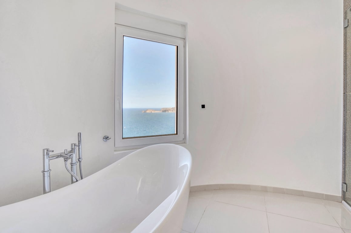 Minimalist bathroom featuring a freestanding white bathtub and chrome fixtures, offering direct views of the Mediterranean Sea through a large window.