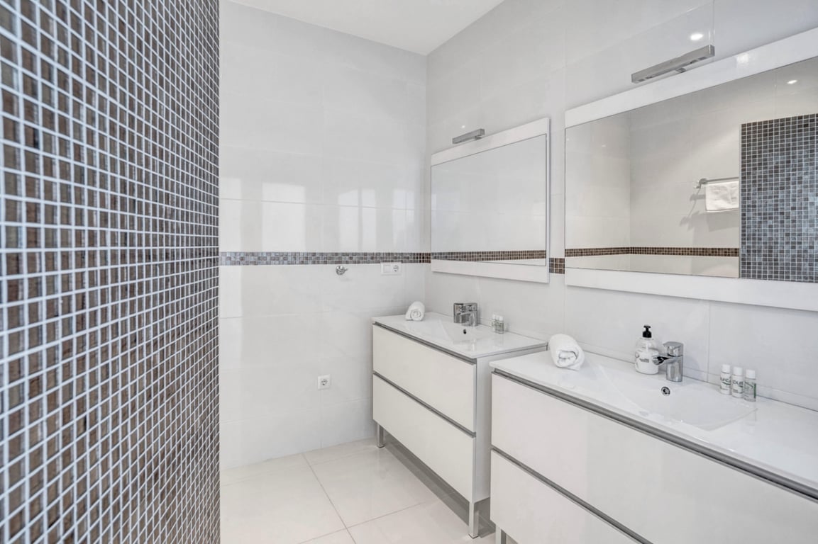 Modern bathroom featuring a double vanity with minimalist white cabinetry, integrated sinks, and dark mosaic tile accents on the walls.