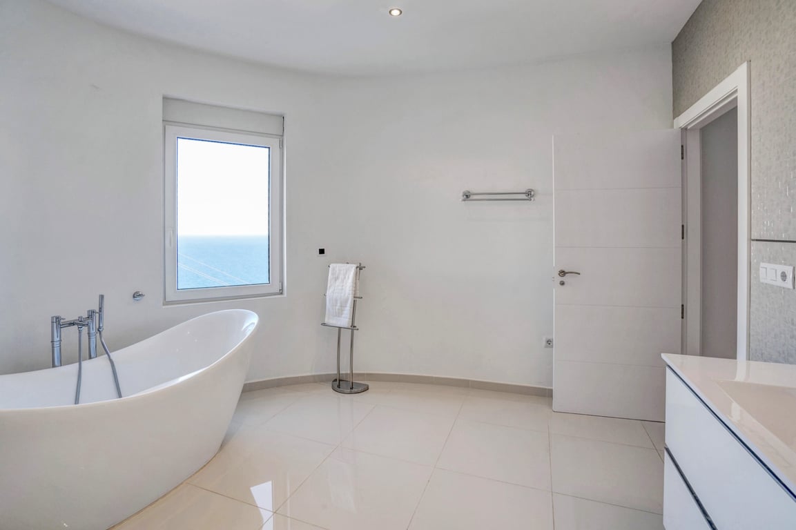 Modern bathroom featuring a freestanding oval bathtub, polished tile flooring, and a window offering views of the Mediterranean horizon.