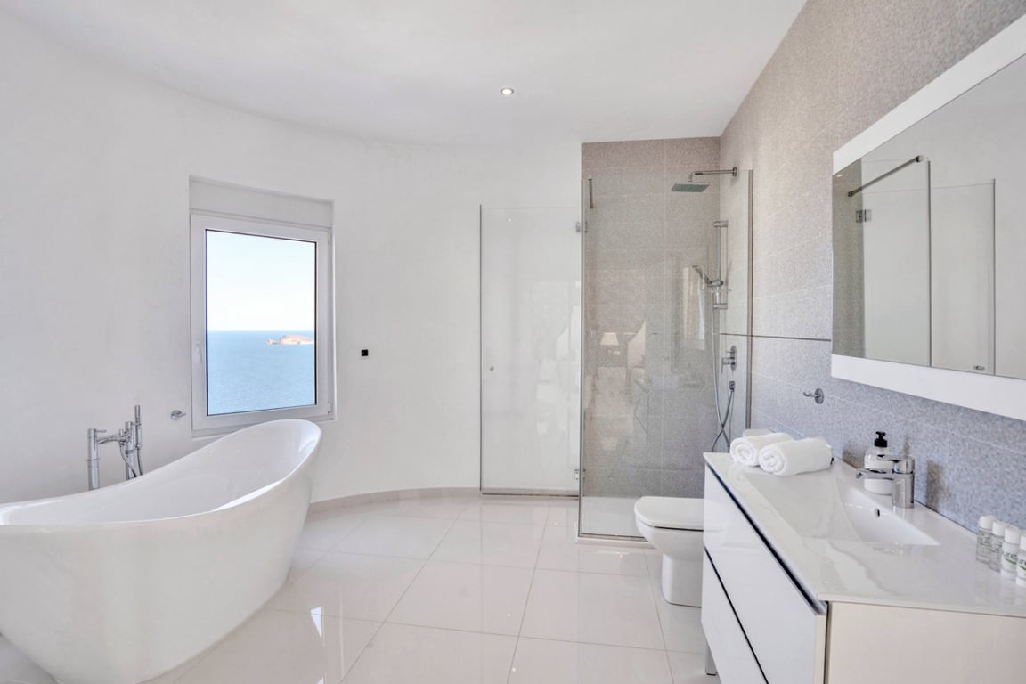 Modern bathroom featuring a freestanding oval bathtub with sea views, a glass-enclosed walk-in shower, and a sleek white vanity with a large mirror.