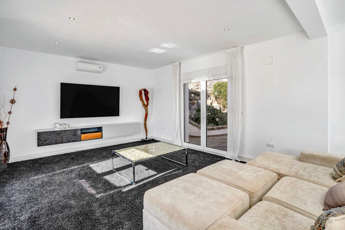 Modern media lounge featuring a modular sofa, dark shag rug, and floating console, with glass doors opening to a terrace bordered by natural rock.