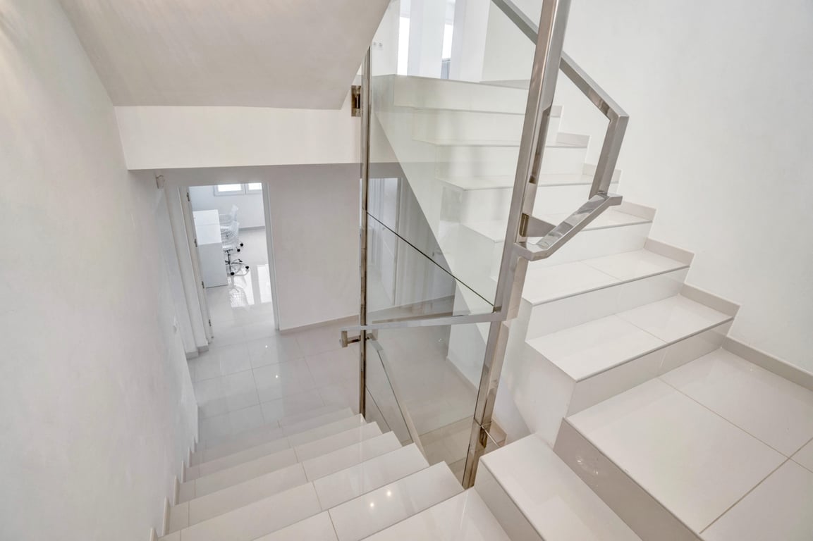 Modern staircase with white tiled steps, glass panels, and stainless steel railings, reflecting a bright and minimalist interior design.