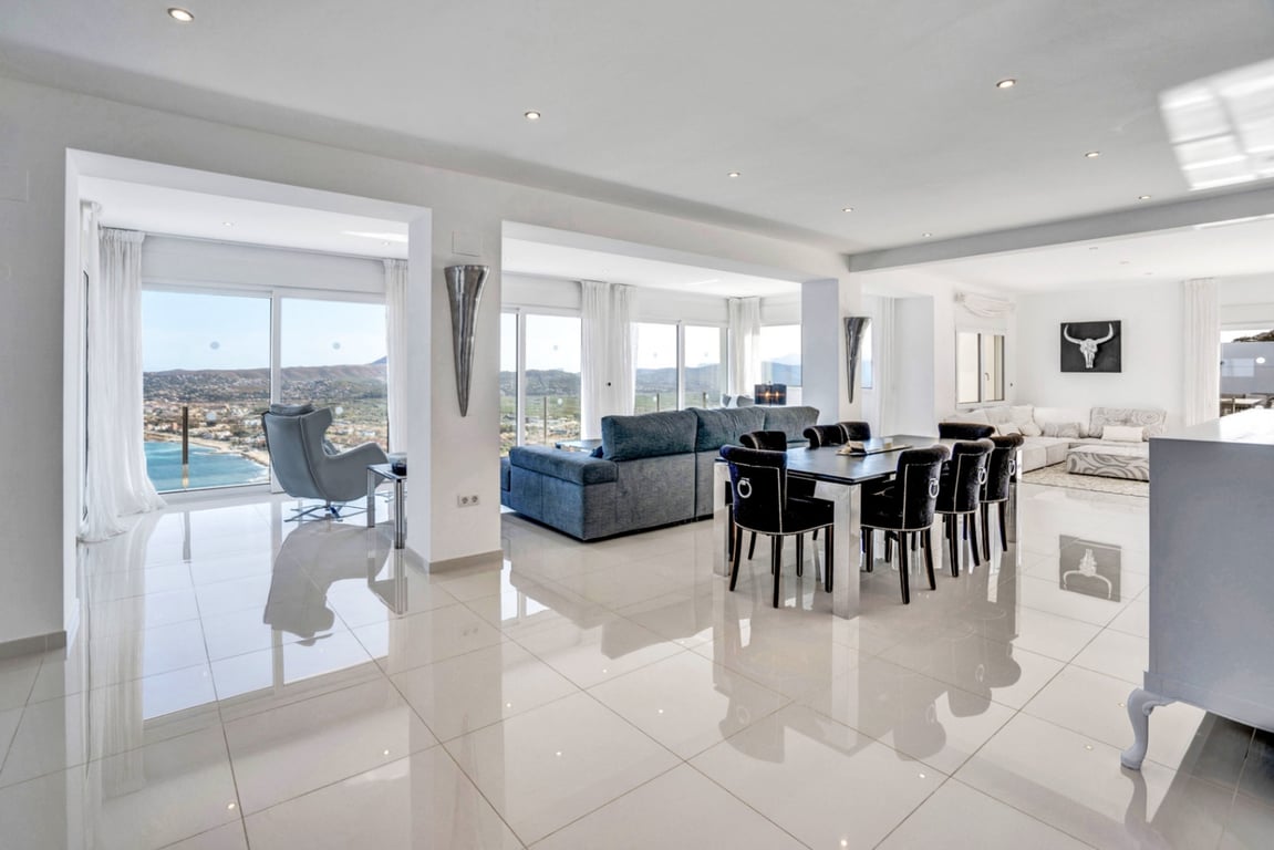 Open-plan living and dining space featuring high-gloss flooring and floor-to-ceiling glass doors with panoramic views of the Mediterranean coast and mountains.