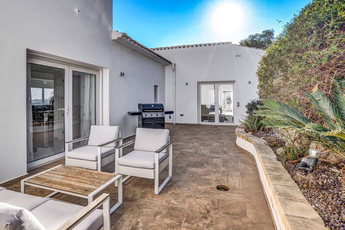 Outdoor terrace featuring stone tiling, modern lounge seating, a gas barbecue, and a curved stone planter with Mediterranean greenery.