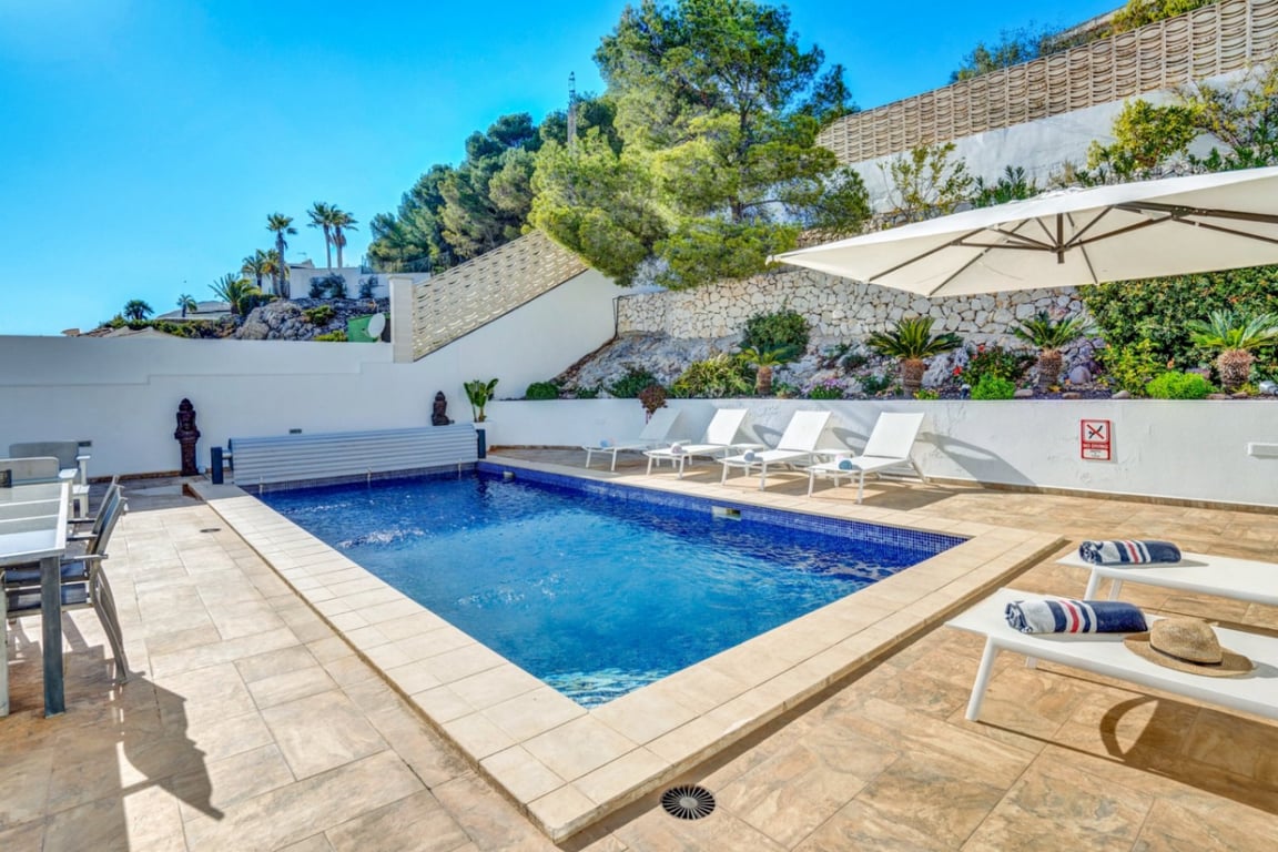 Private outdoor pool featuring a blue mosaic finish, stone-tiled sun deck with white loungers, and a natural stone wall with Mediterranean vegetation.
