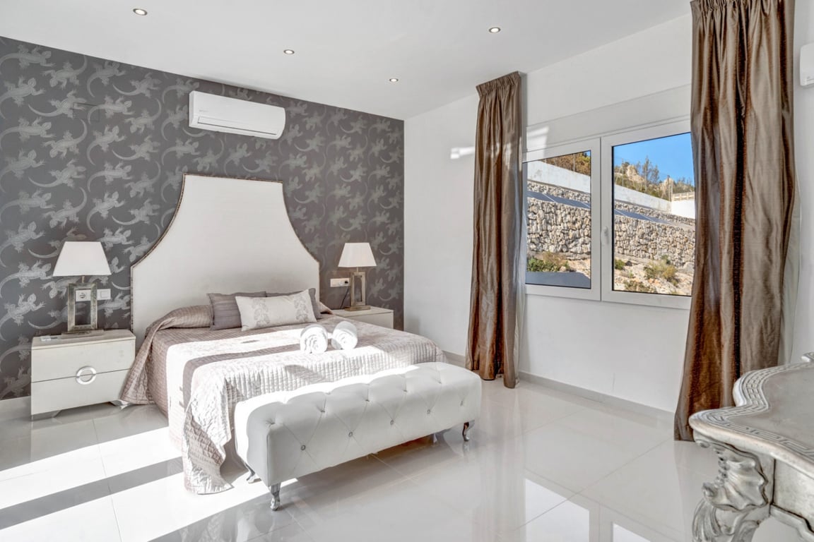 Suite bedroom featuring a grey patterned accent wall, white tiled flooring, air conditioning, and a window overlooking the natural stone landscape.