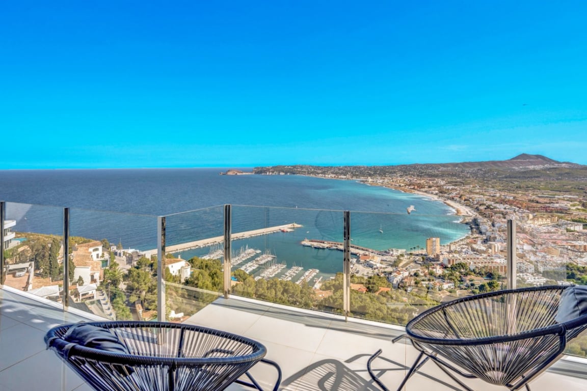 Terrace featuring glass railings and black Acapulco chairs, providing panoramic views over the Jávea port and the Mediterranean coastline.
